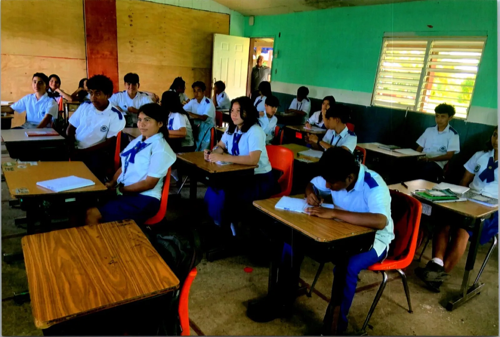Secondary students in white and blue uniforms seated at wooden desks in a classroom