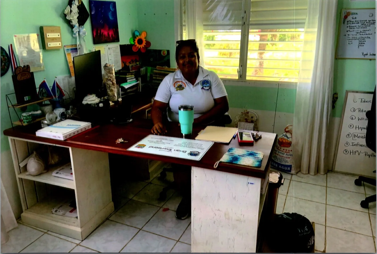 A staff member seated at a desk in a school office with green walls and louvred windows