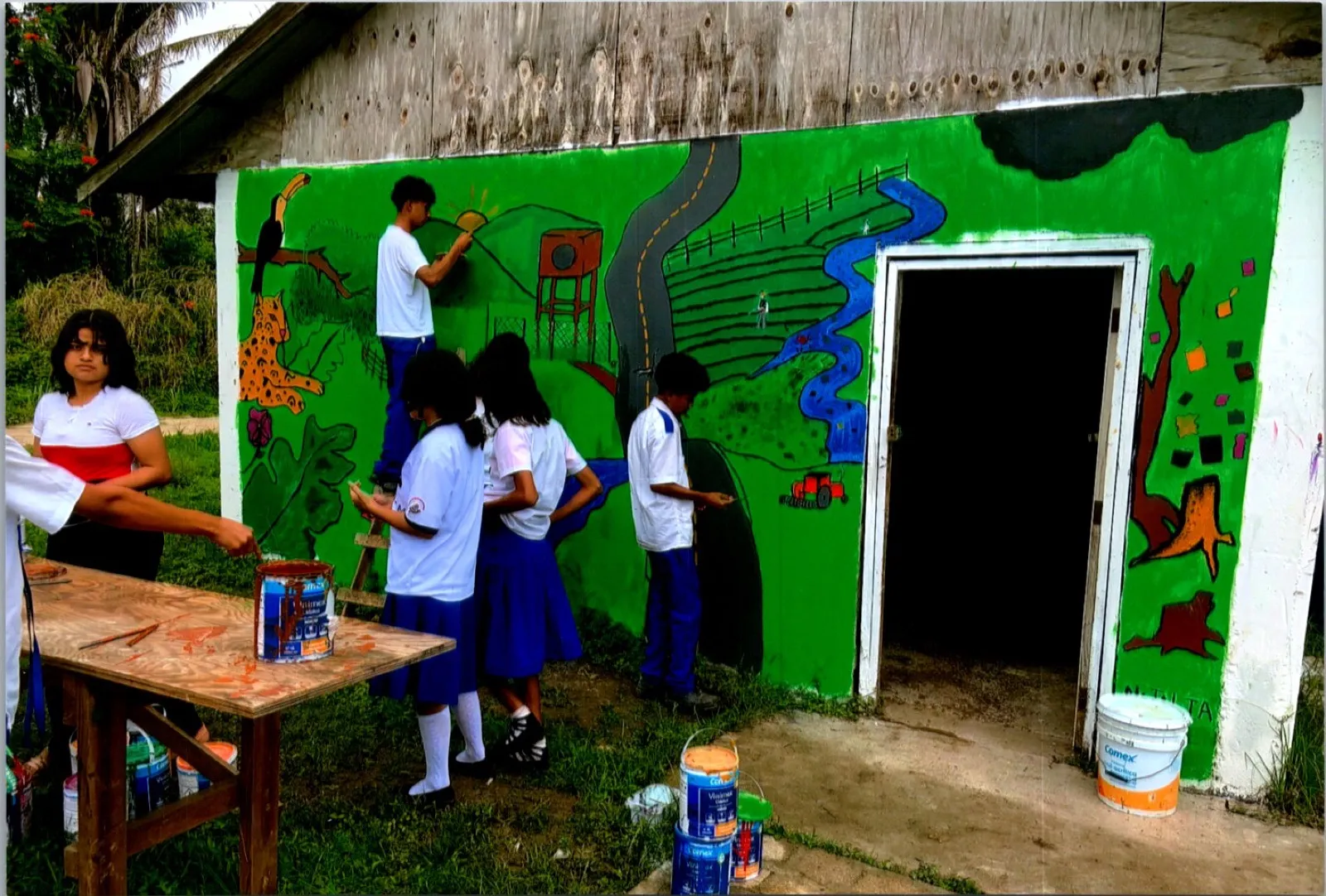 Students in school uniforms painting a green mural of the countryside on a school building in the Valley of Peace, Belize